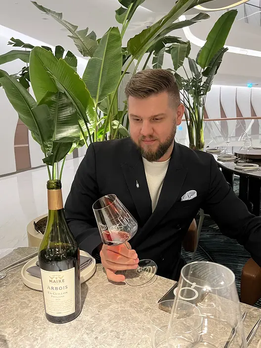 Alex Kovalov tasting a glass of Jura wine at a restaurant table, a bottle of Arbois beside him